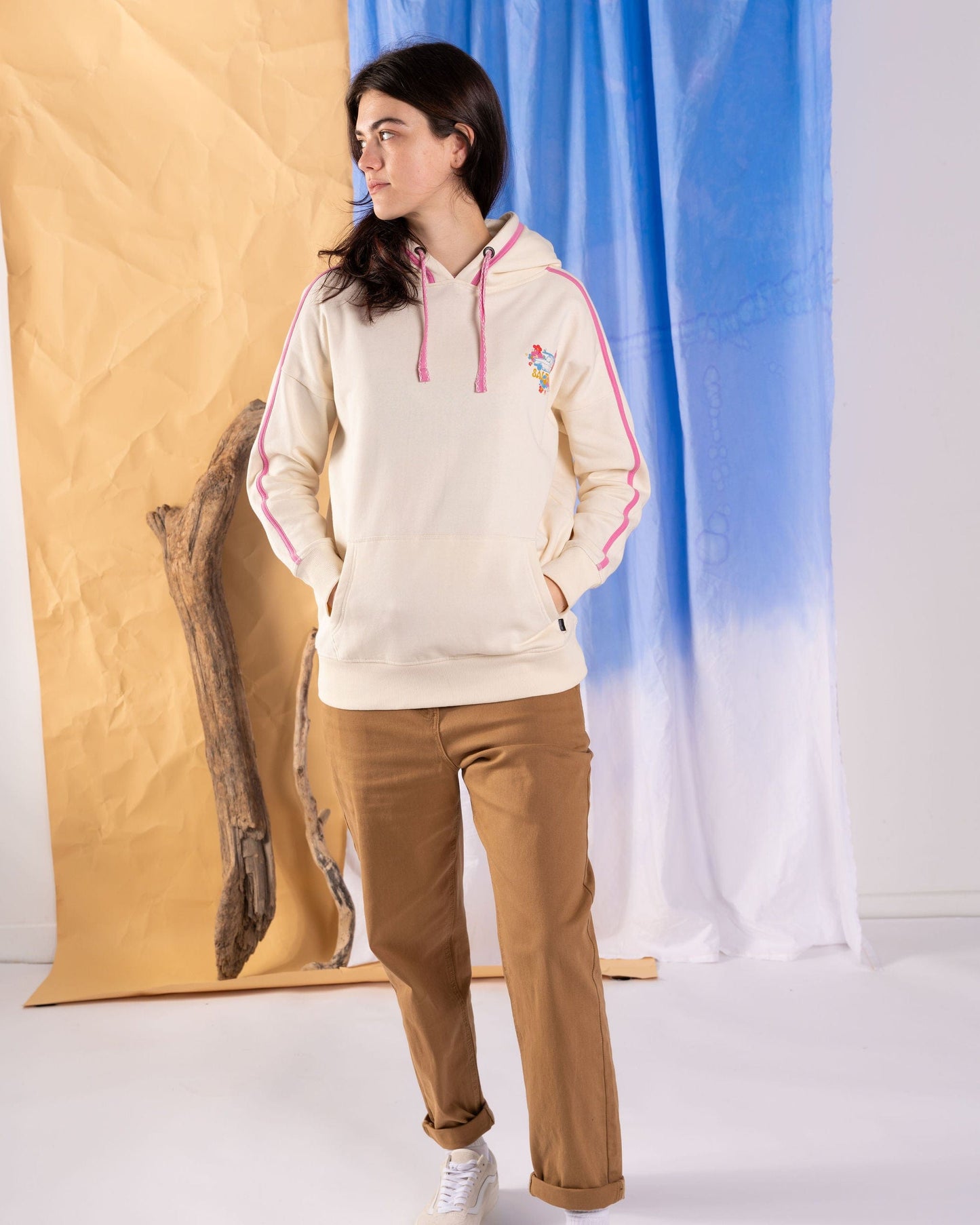 Wearing Saltrock's "Highway" Women's Pop Hoodie in cream with pink contrast details and a kangaroo pocket, a person stands indoors near a textured blue and beige fabric backdrop. Their brown pants perfectly complement Saltrock's signature style.