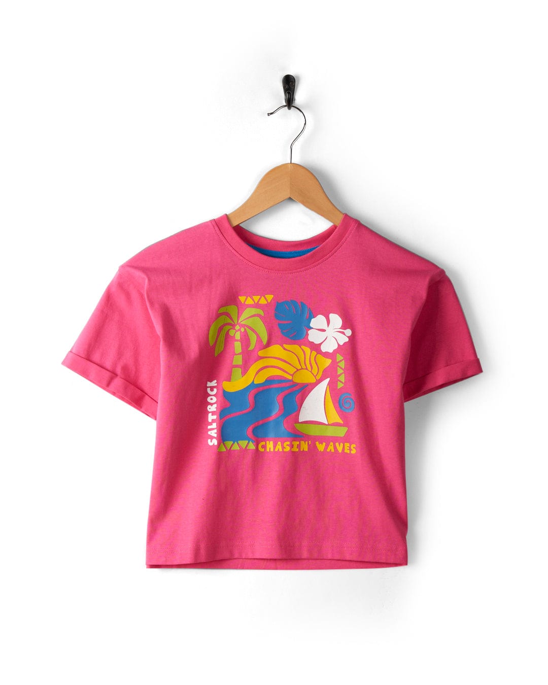 Summer Block - Kids Recycled Cropped T-Shirt - Pink with colorful Saltrock graphics, featuring a palm tree, sun, waves, and a sailboat. The text reads "Sunsick" and "Chasin' Waves." Made from recycled materials for an eco-friendly choice. The cropped boxy fit t-shirt is hanging on a wooden hanger.