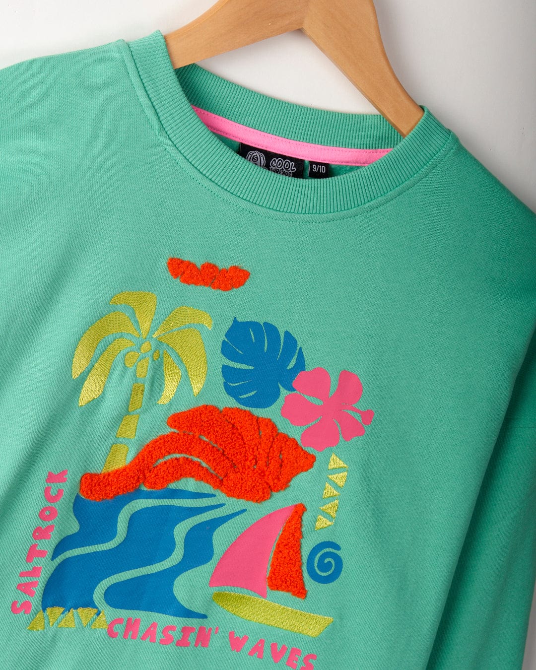 Green cropped-fit sweatshirt on a wooden hanger with vibrant beach-themed embroidered graphics. Featuring palm trees, waves, a sailboat, and the text "Saltrock" and "Chasin' Waves," the Summer Block - Kids Sweat - Green from Saltrock is perfect for those who love Saltrock branding and coastal vibes.