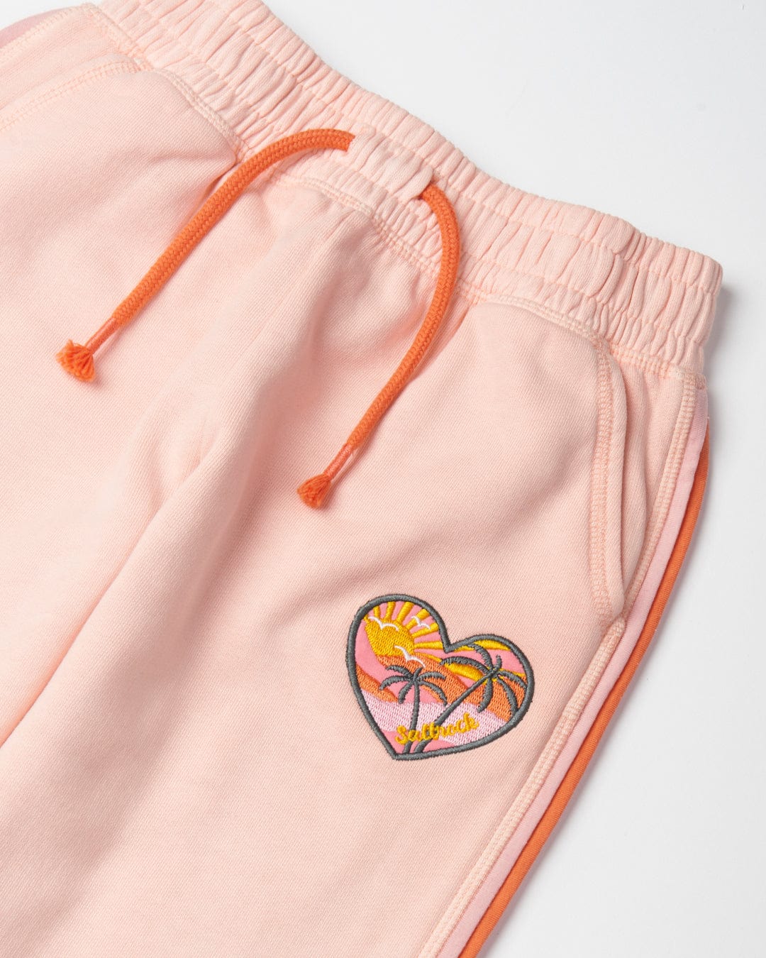 Close-up of Sunshine State - Kids Joggers - Peach with an orange drawstring and an embroidered heart patch. The patch features a sunset with palm trees and the word "sunset." Made from 100% cotton, these joggers from Saltrock offer a soft hand feel and have an elasticated waistband for ultimate comfort.