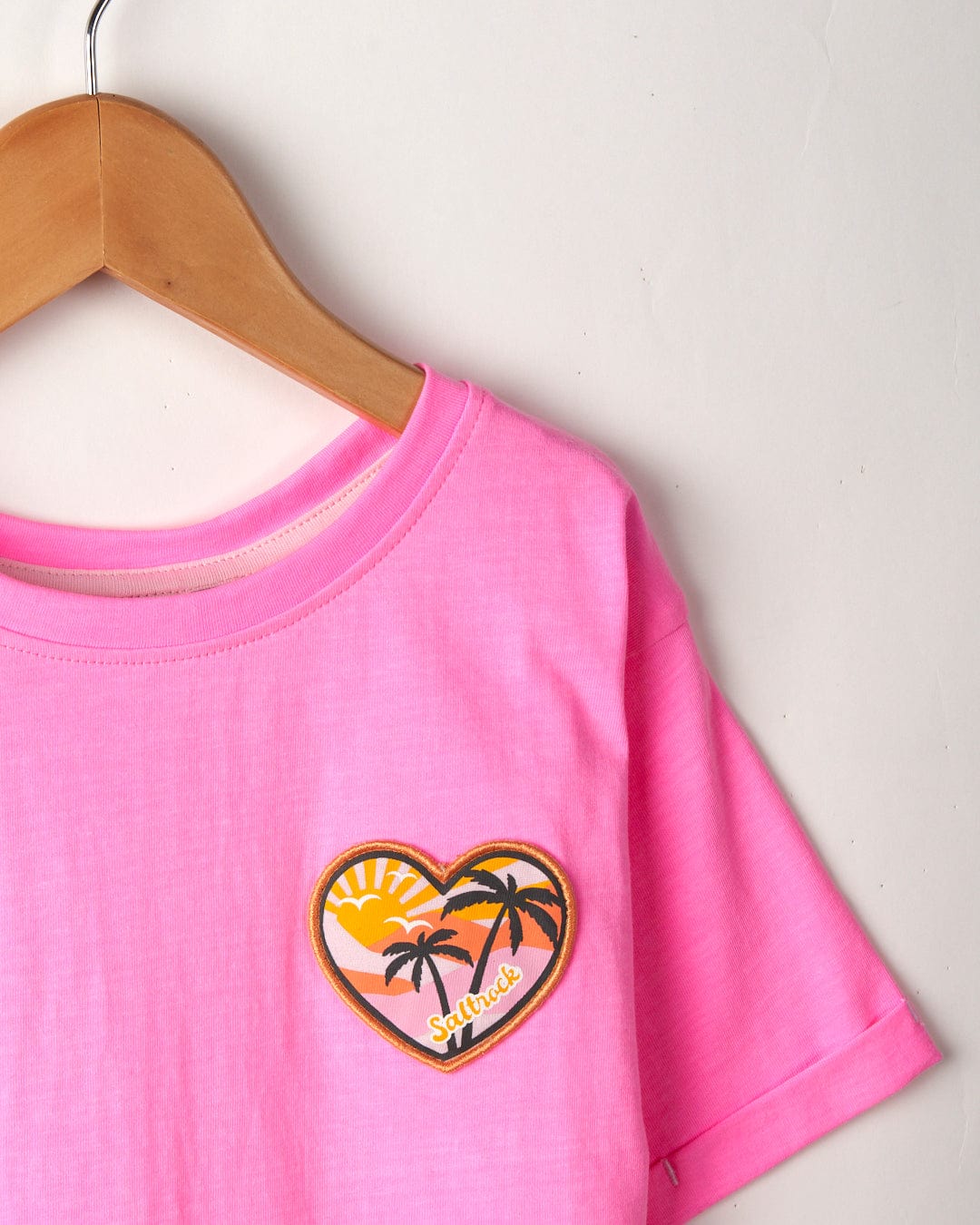 Sunshine State - Recycled Kids T-Shirt - Pink by Saltrock, with a tropical island-themed heart patch, hanging on a wooden hanger against a white wall.