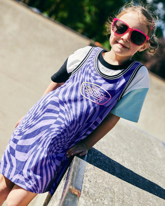 A young child wearing pink sunglasses and a **Saltrock Rezz - Kids Recycled Warp Vest Dress - Purple** stands outdoors, leaning on a concrete barrier on a sunny day.