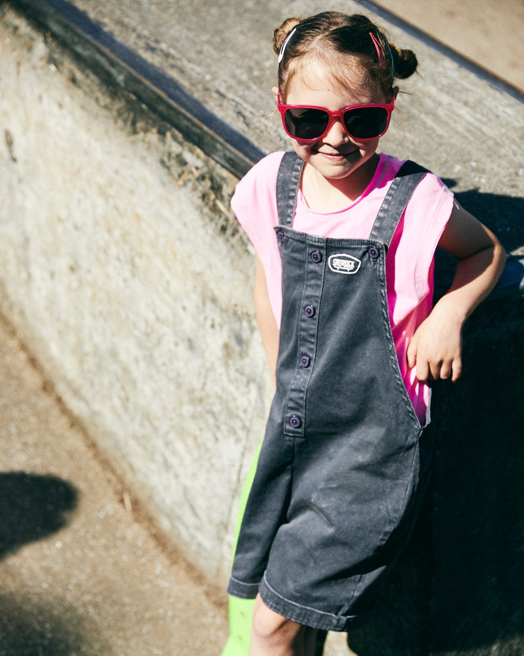 A young child wearing red sunglasses, a Saltrock Mystic Skulls - Recycled Kids Longline Vest - Pink, and denim overalls leans against a concrete wall outdoors, exuding a distressed grunge style.