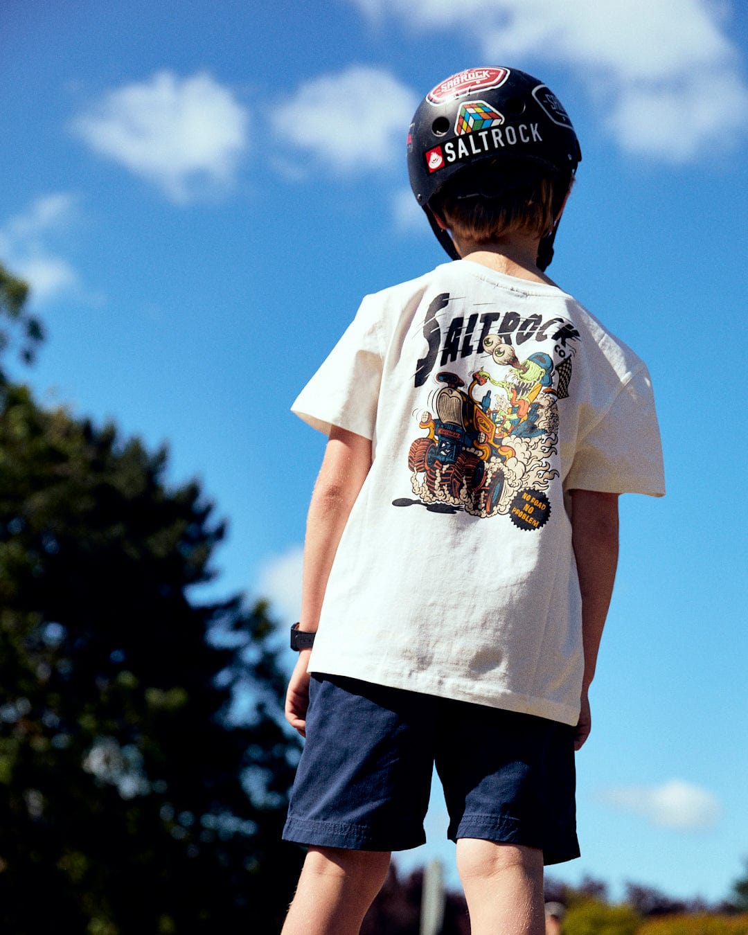 A child wearing a helmet and a Saltrock No Road No Problem - Kids Short Sleeve T-Shirt - White with vibrant Saltrock illustrations stands outdoors under a blue sky.