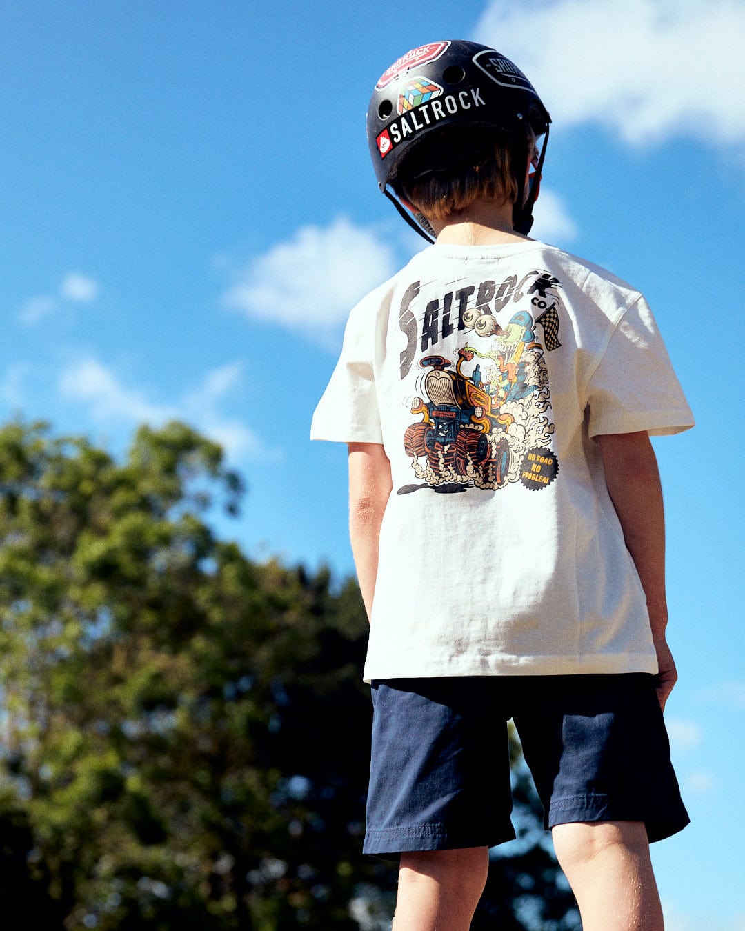 A child wearing a helmet and a t-shirt with Saltrock illustrations stands outdoors, viewed from the back, with tree branches and a blue sky visible above, wearing the No Road No Problem - Kids Short Sleeve T-Shirt - White by Saltrock.