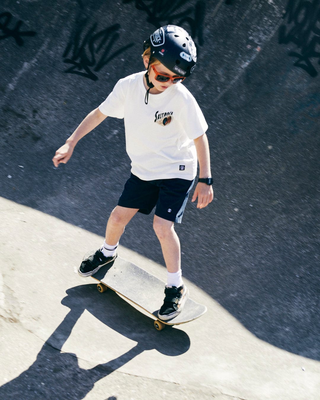 A young boy, wearing a helmet, sunglasses, a white No Road No Problem - Kids Short Sleeve T-Shirt - White from Saltrock that's machine washable, and shorts, skateboards in a skate park.
