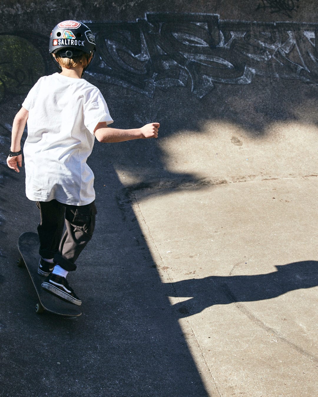 A person wearing a helmet and a Taco Tok - Kids Short Sleeve T-Shirt by Saltrock is skateboarding on a concrete surface at a skate park with graffiti.