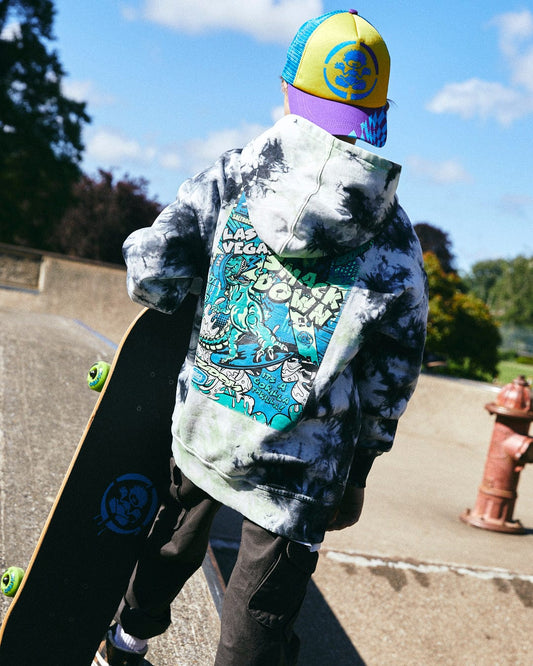 A person wearing a Saltrock Las Vegas Smackdown - Kids Glow in the Dark Oversized Pop Hoodie - Multi and colorful cap holds a skateboard while standing on a concrete surface. Trees and a fire hydrant are in the background.