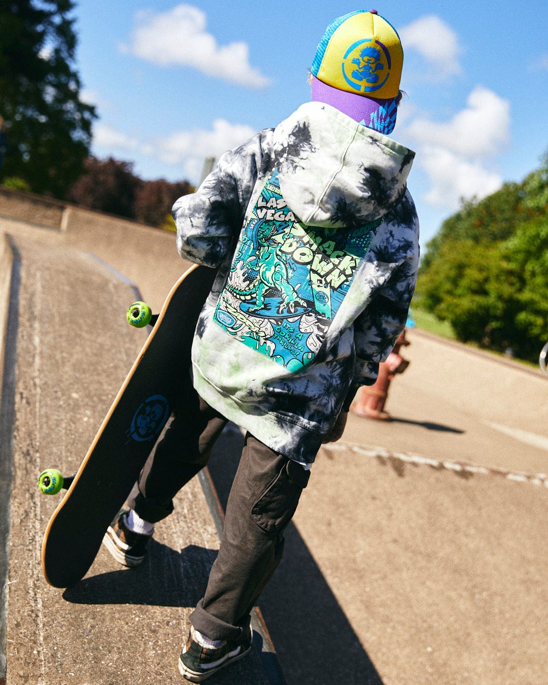 A person dressed in a Saltrock Las Vegas Smackdown - Kids Glow in the Dark Oversized Pop Hoodie - Multi and colorful hat holds a skateboard while standing at a skate park.