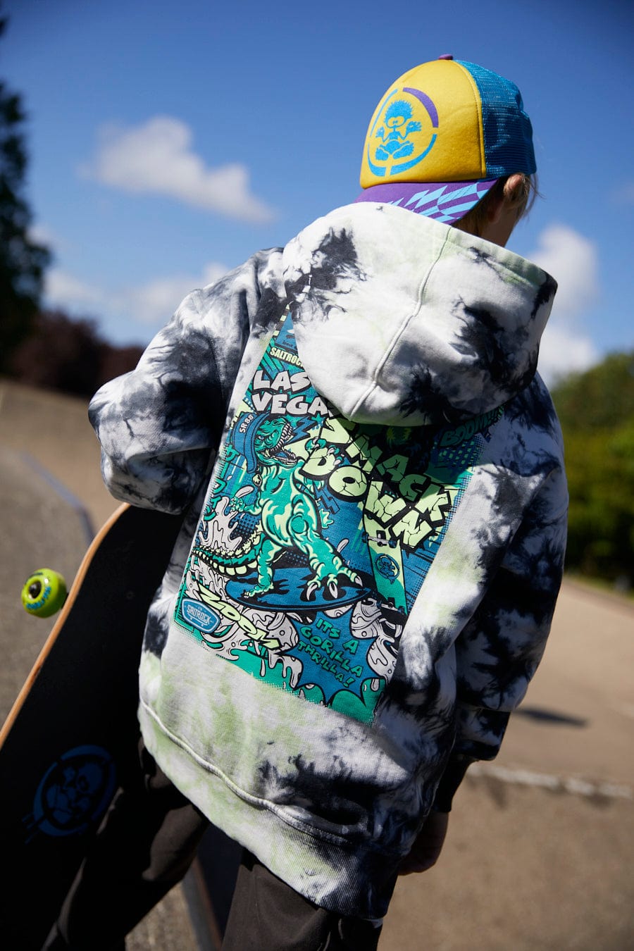Person wearing a multi-colored Saltrock cap and a Saltrock Las Vegas Smackdown - Kids Glow in the Dark Oversized Pop Hoodie - Multi with a comic-style graphic on the back, holding a skateboard, standing outdoors.