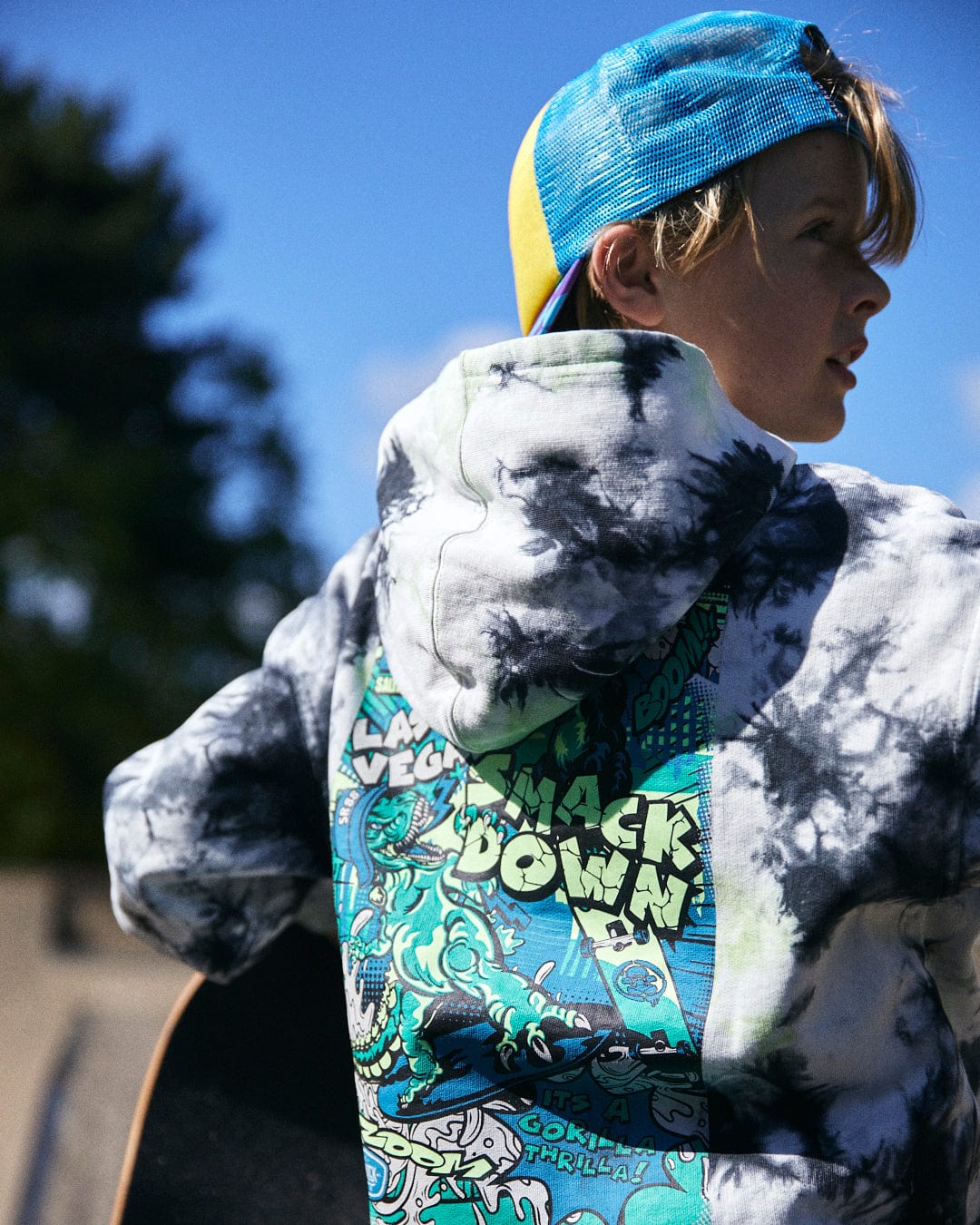 A person wearing a Saltrock Las Vegas Smackdown - Kids Glow in the Dark Oversized Pop Hoodie - Multi with a colorful graphic print and a blue and yellow cap, holding a skateboard. Trees and a blue sky are in the background.