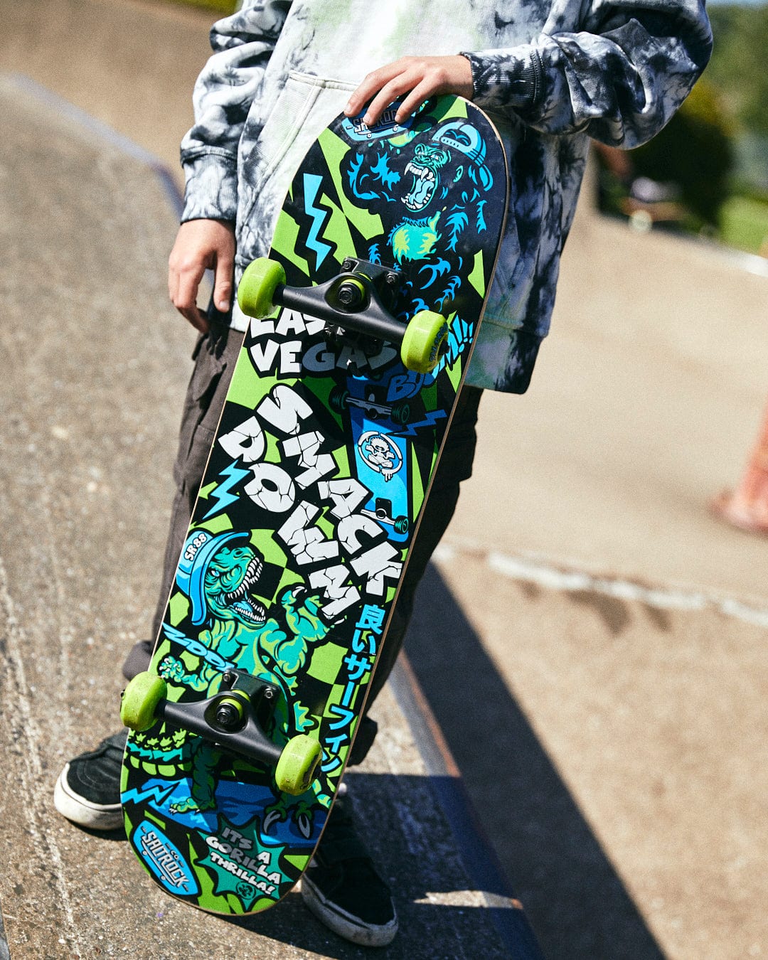 A person wearing a tie-dye hoodie holds a colorful, hardwearing Saltrock Las Vegas Smackdown - Skateboard - Green/Blue with green wheels and graphic designs, including the words "SMACK DOWN" and various illustrations.