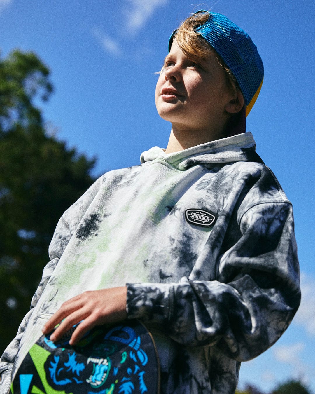 A person wearing a tie dye Saltrock Las Vegas Smackdown - Kids Glow in the Dark Oversized Pop Hoodie - Multi and a backward cap holds a skateboard. They are looking off into the distance with trees and a blue sky in the background.