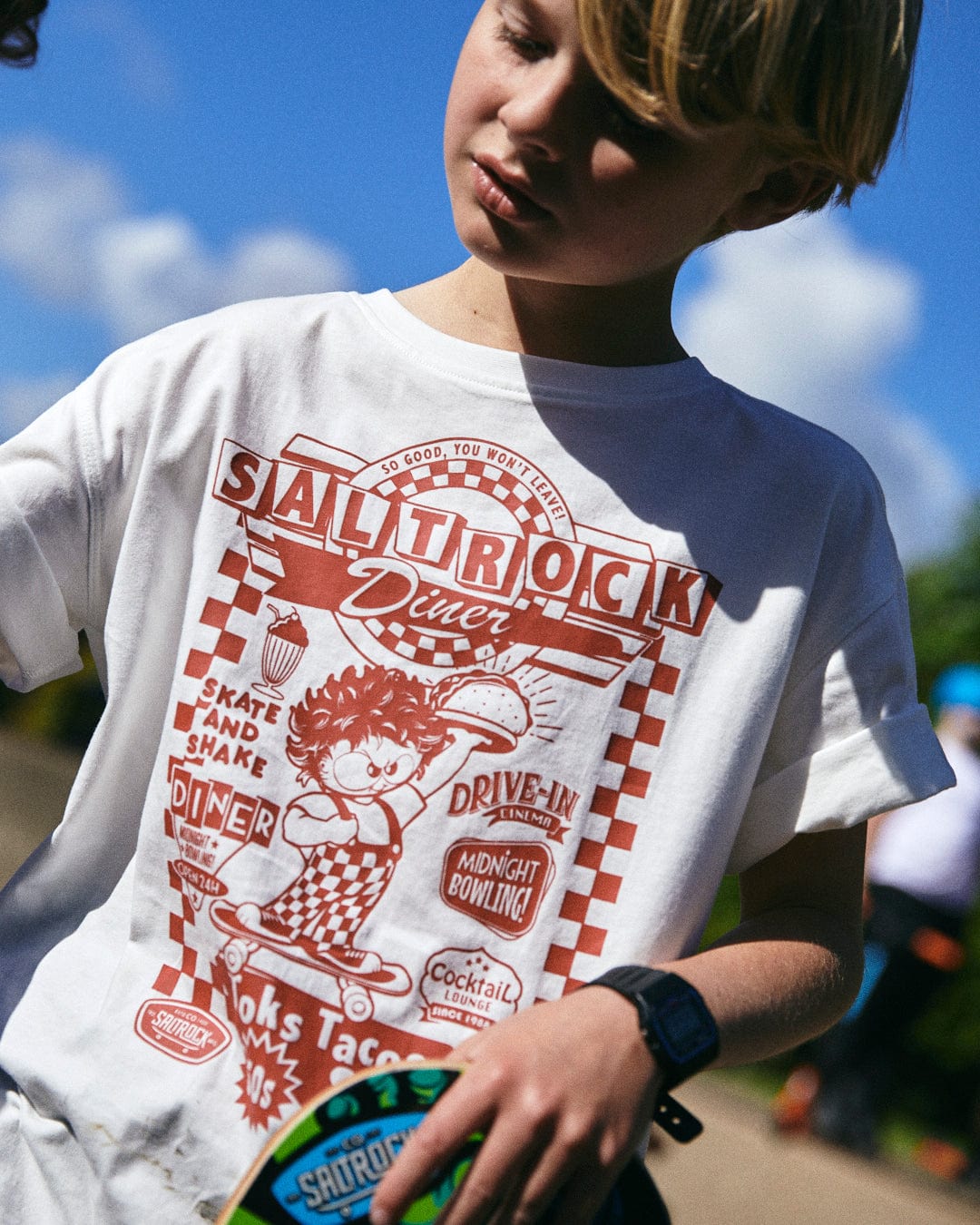 A young child wearing a white "Taco Tok - Kids Short Sleeve T-Shirt" by Saltrock, made of 100% Cotton, is holding a skateboard. The t-shirt features a retro-style design with various diner-themed illustrations and text.