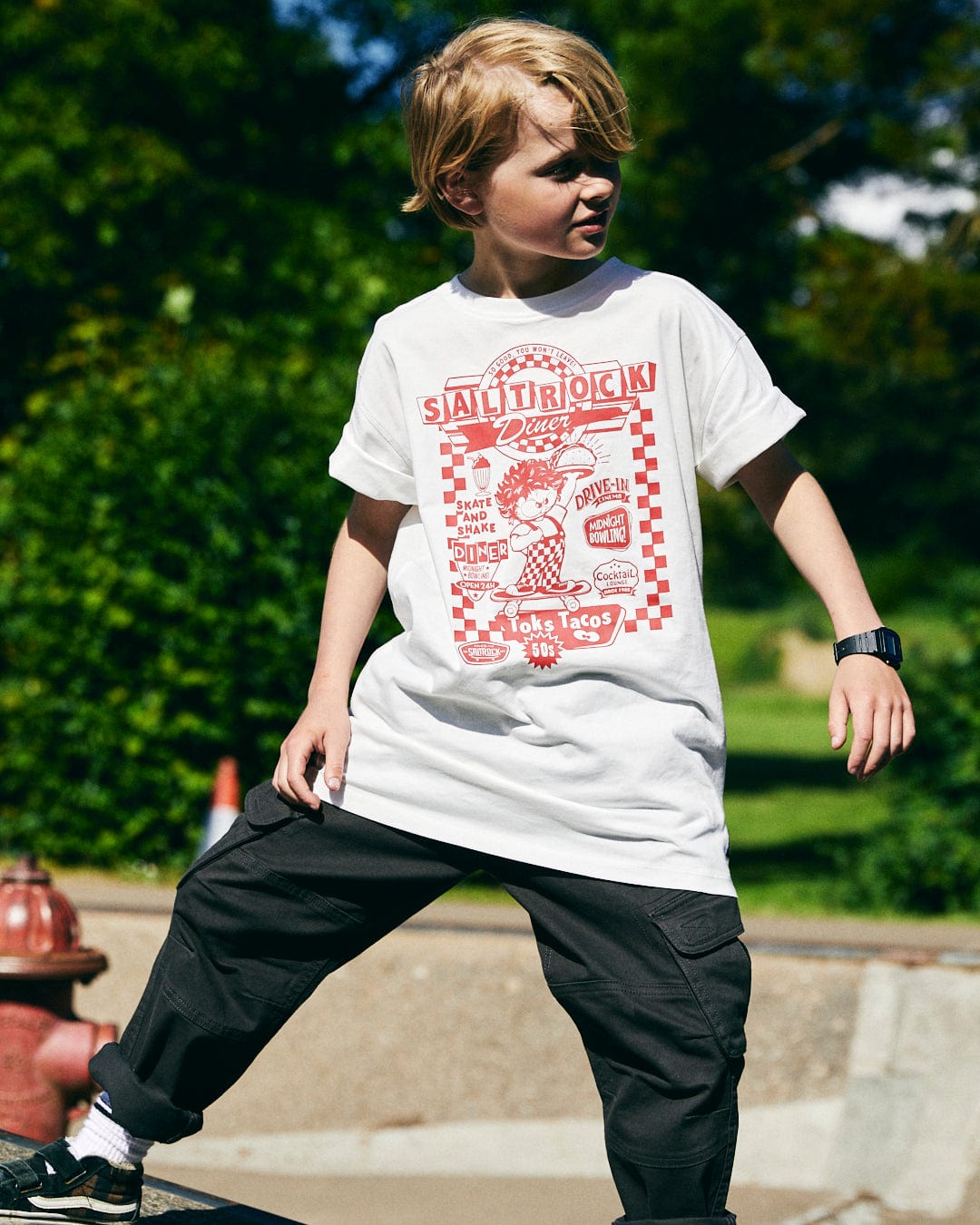 A young boy with blond hair stands outdoors wearing a white T-shirt featuring the "Taco Tok - Kids Short Sleeve T-Shirt" by Saltrock and black pants. He is looking to the side with one foot on a slightly higher surface.
