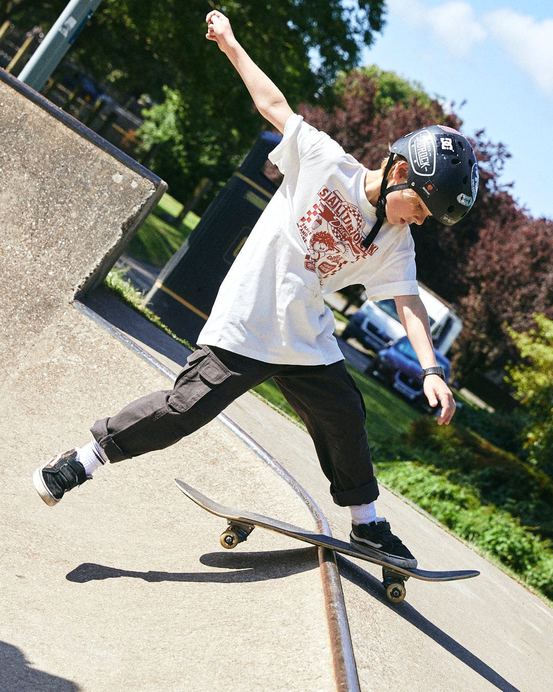 A child wearing a helmet and protective gear skateboards on a rail at a skate park, showcasing their Saltrock Taco Tok - Kids Short Sleeve T-Shirt logo, surrounded by green trees and a sunny sky.