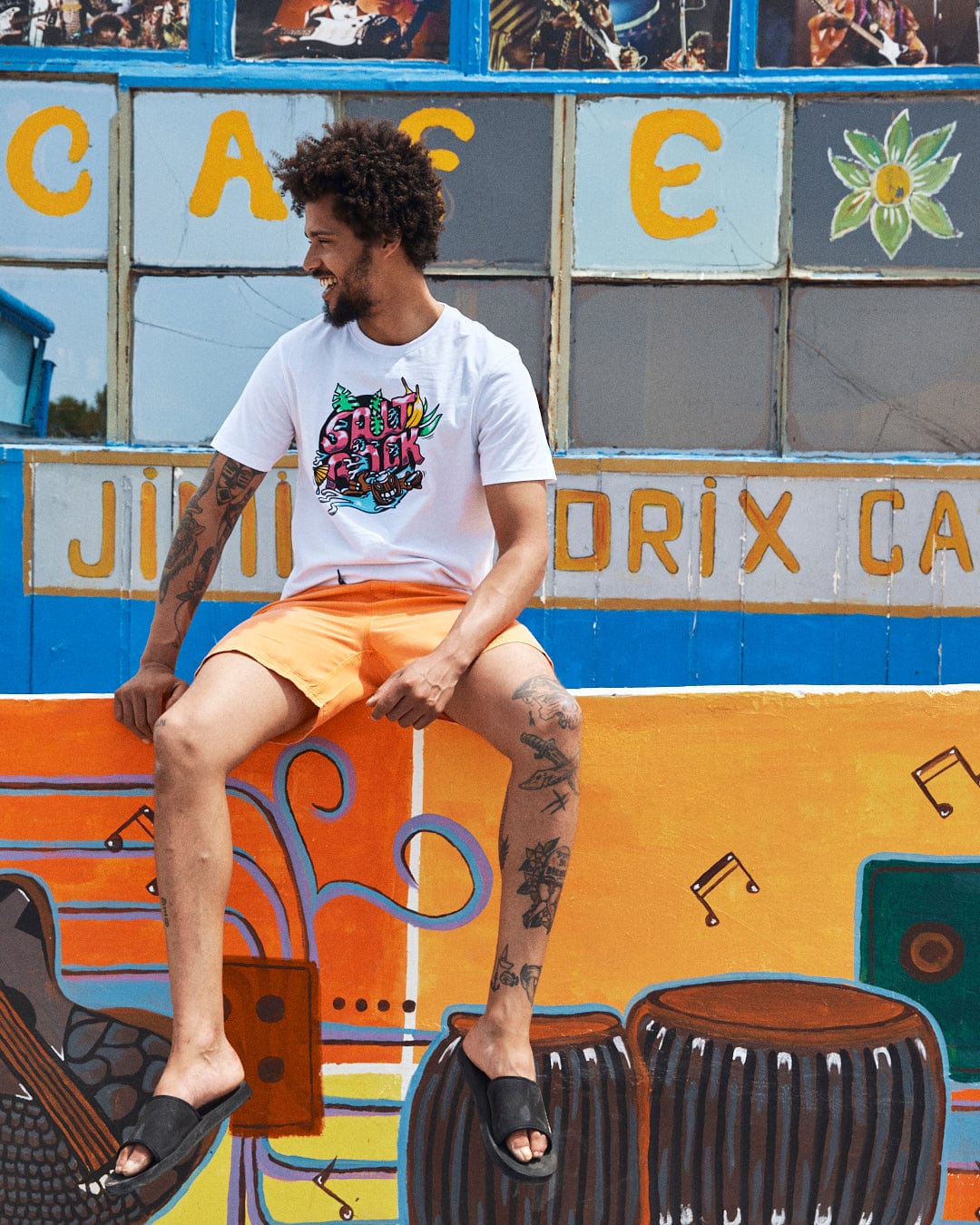 A man with tattoos, wearing a 100% cotton white Tahiti - Mens Short Sleeve T-Shirt by Saltrock and orange shorts, sits on a colorful mural featuring music notes, drums, and the name "Jimi Hendrix Café," showcasing a vibrant Saltrock illustration.