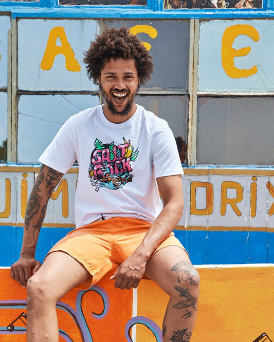 A person with curly hair and tattoos sits smiling in front of a colorful building. They are wearing a white Saltrock Tahiti - Mens Short Sleeve T-Shirt - White made from 100% cotton and orange shorts.