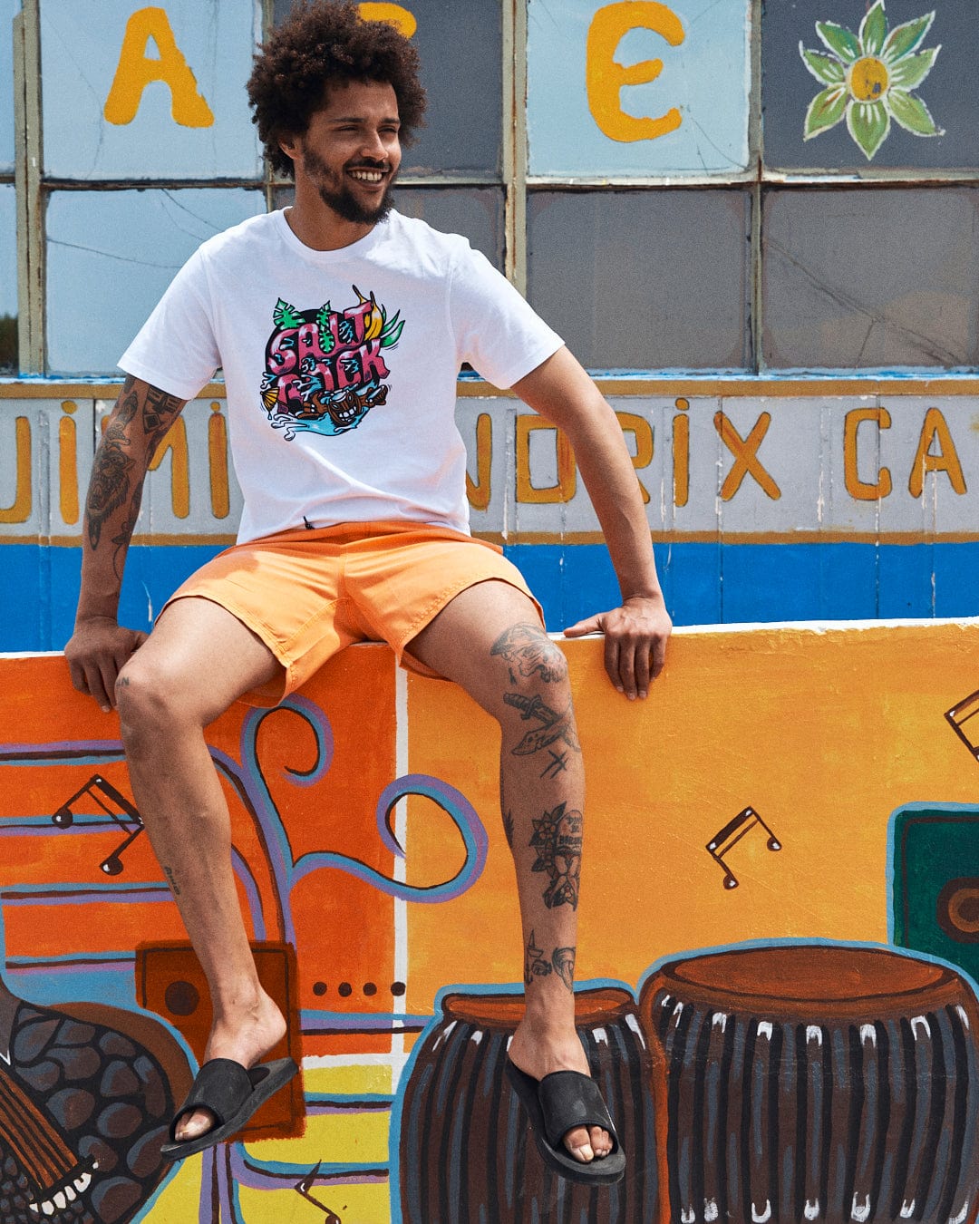 A man with tattoos, wearing a 100% cotton, illustrated white Tahiti - Mens Short Sleeve T-Shirt - White by Saltrock and orange shorts, sits on a colorful wall mural. He appears to be smiling, looking slightly left.