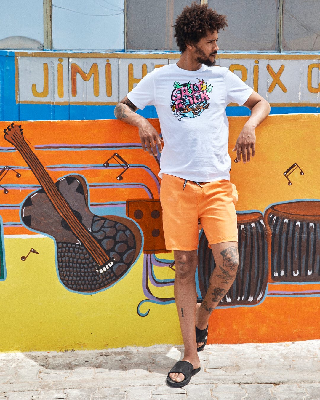 A man in a Saltrock Tahiti - Mens Short Sleeve T-Shirt - White and orange shorts stands in front of a brightly colored mural featuring musical instruments, resembling a vibrant Saltrock illustration.