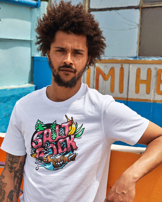 Man with curly hair wearing a 100% cotton white Tahiti - Mens Short Sleeve T-Shirt - White featuring a colorful Saltrock illustration poses outdoors near vibrant walls.