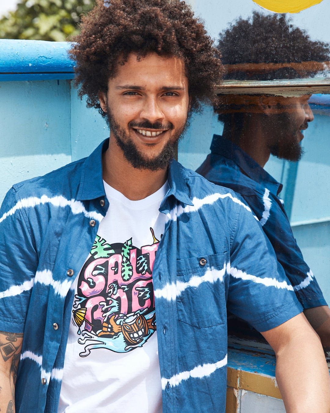 A bearded man with curly hair smiles while wearing a blue tie-dye shirt over a white graphic T-shirt featuring a Tahiti - Mens Short Sleeve T-Shirt - White by Saltrock, standing next to a blue wall with his reflection visible in a mirrored window.