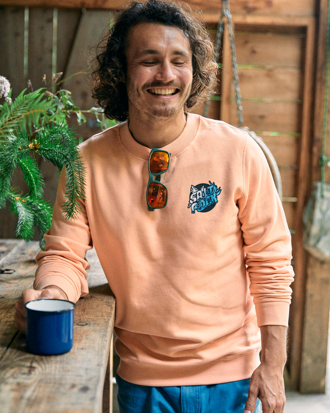 A man with wavy hair and a mustache is smiling while leaning against a wooden table, holding a blue mug. He’s wearing the Saltrock Tahiti Men's Sweat in peach, styled in an oversized fit, with sunglasses hanging from the collar.