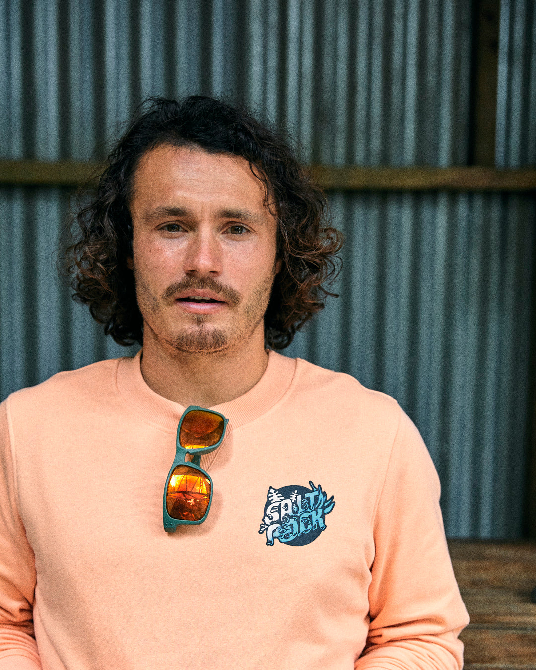 A man with curly hair and a mustache wearing an oversized Saltrock Tahiti peach-colored sweatshirt stands in front of a corrugated metal wall. Sunglasses are hanging on the neckline of his 100% cotton shirt adorned with Saltrock graphics.