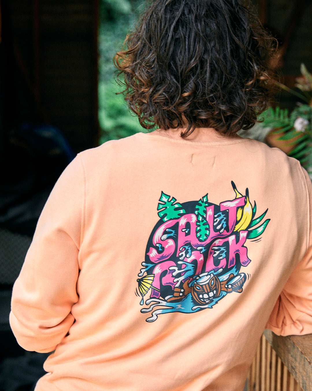 A person with curly hair is wearing an oversized peach-colored Tahiti sweatshirt from Saltrock, made of 100% cotton, featuring vibrant Saltrock graphics on the back that read "Salt Rock" alongside illustrations of tropical elements.