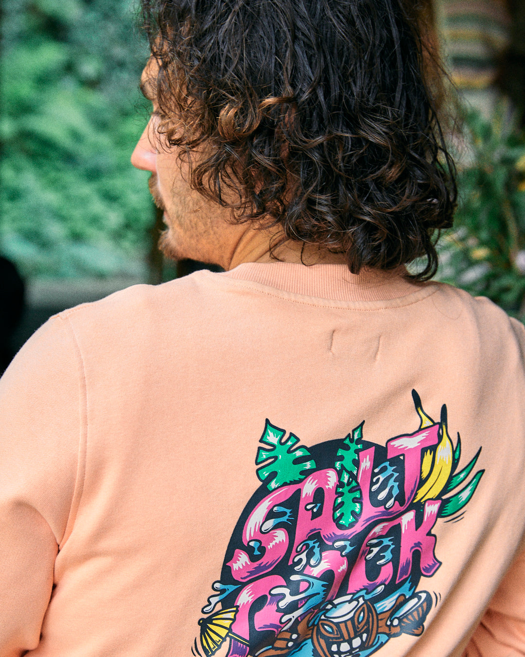 A person with curly hair is seen from the back, wearing the "Tahiti - Mens Sweat - Peach" by Saltrock, featuring a vibrant graphic design that includes the text "Saltrock" and various colorful elements. The shirt is made from 100% cotton and has an oversized fit.