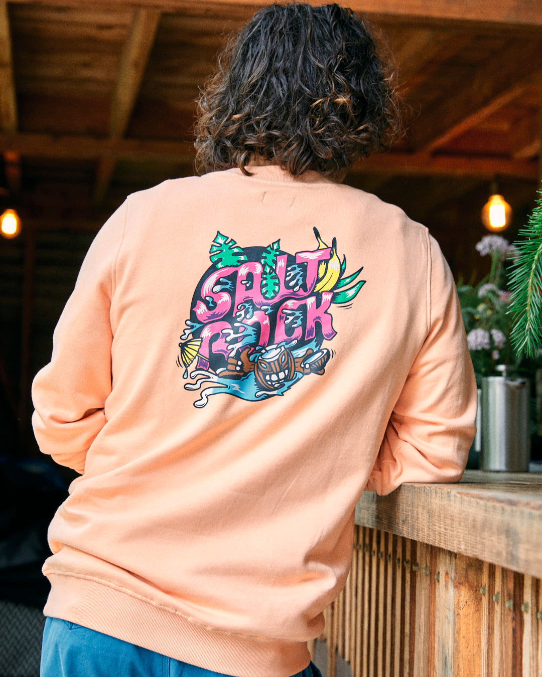 Person in a Tahiti men's sweat in peach, featuring an authentic Saltrock illustration and the text "Saltpack" on the back, standing beside a wooden counter.