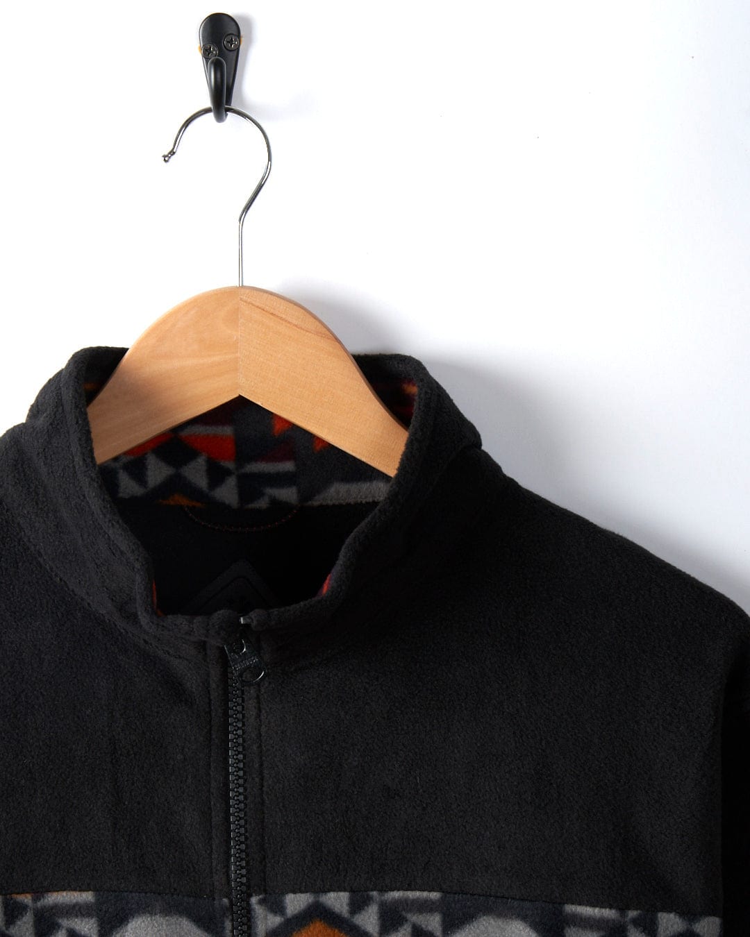 The Theo Wilds - Mens Recycled Microfleece in black by Saltrock hangs on a wooden hanger against a white wall, showcasing a geometric print along the inner collar and near the zipper. A black metal hook supports the hanger.