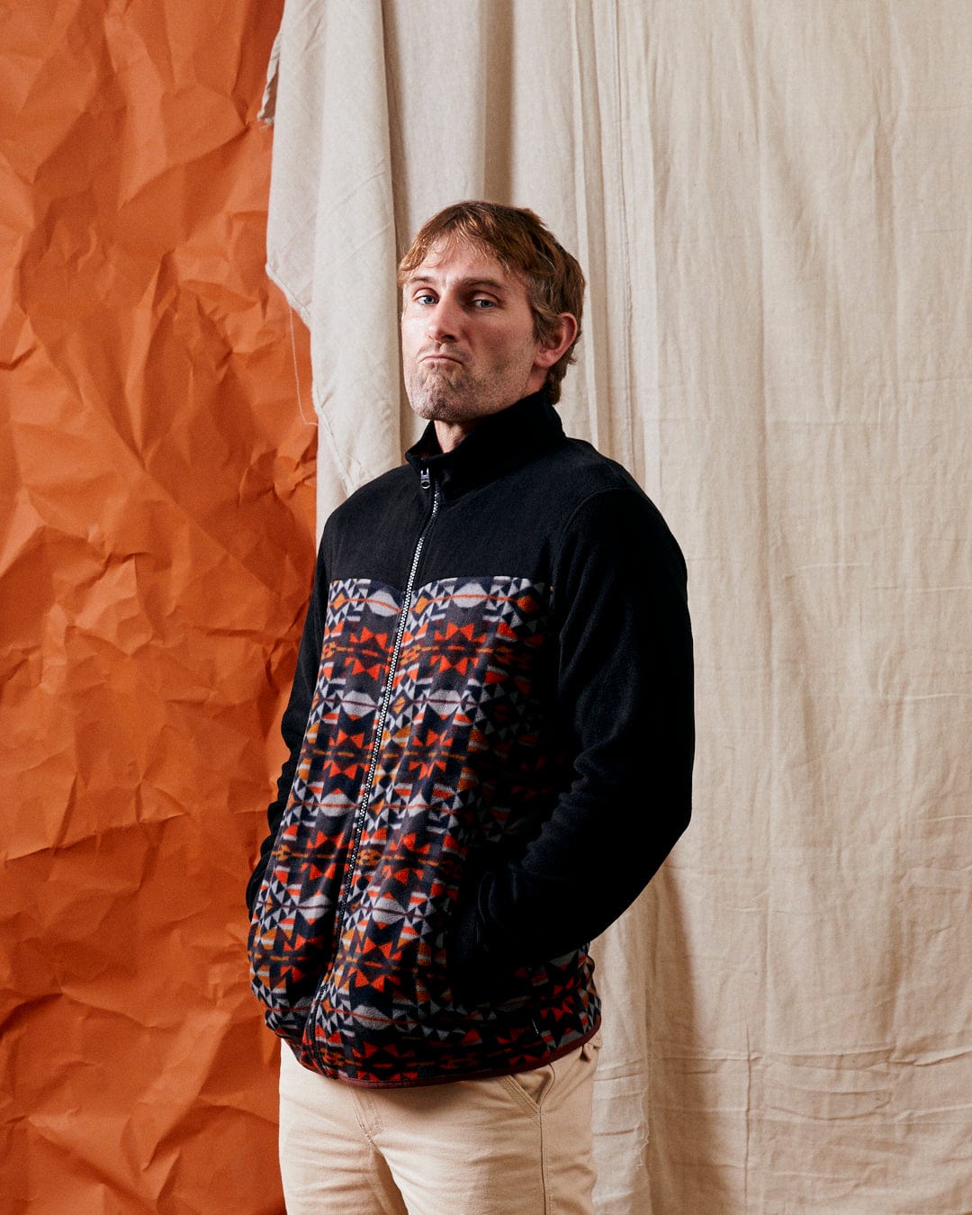 A man wearing the Theo Wilds Men's Recycled Microfleece Jacket in black by Saltrock, with his hands in his pockets, stands in front of a backdrop of crumpled orange and beige fabric.