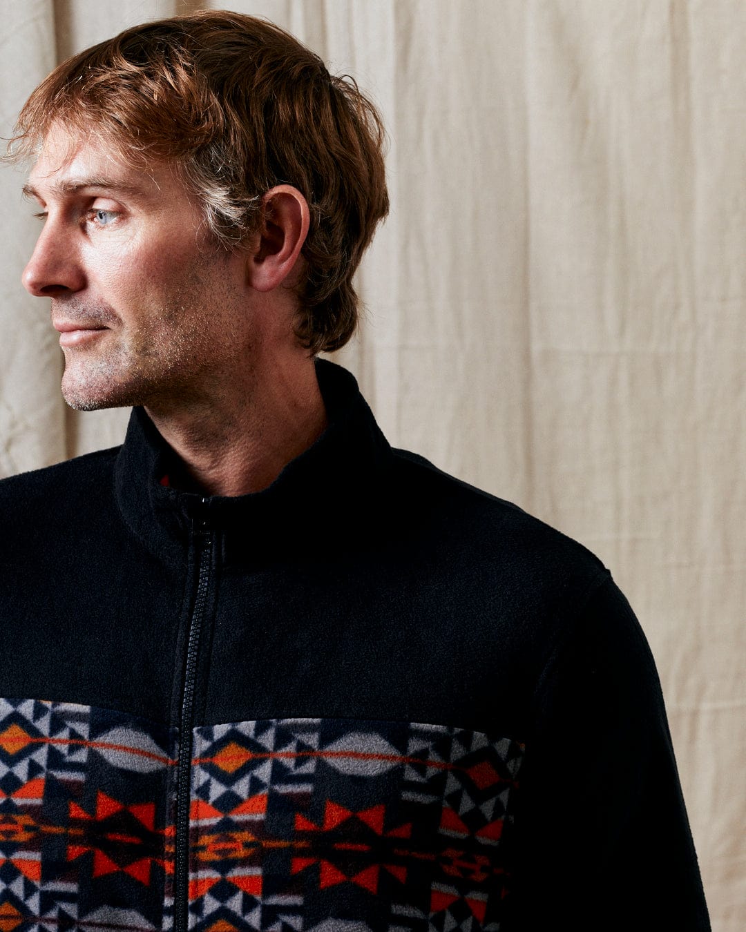 A man with short brown hair wearing the Saltrock Theo Wilds Men's Recycled Microfleece in black, featuring a colorful geometric print, looks to his left while standing against a cream-colored fabric background.