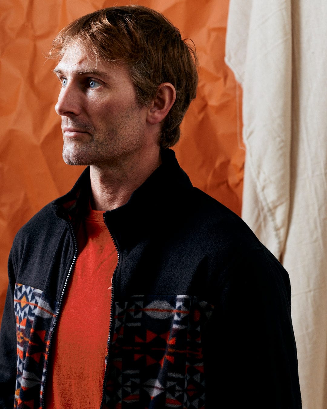 A man with light brown hair wearing the Theo Wilds Men's Recycled Microfleece in black by Saltrock, adorned with a geometric print over an orange shirt, poses against a backdrop of orange and beige textured fabric.