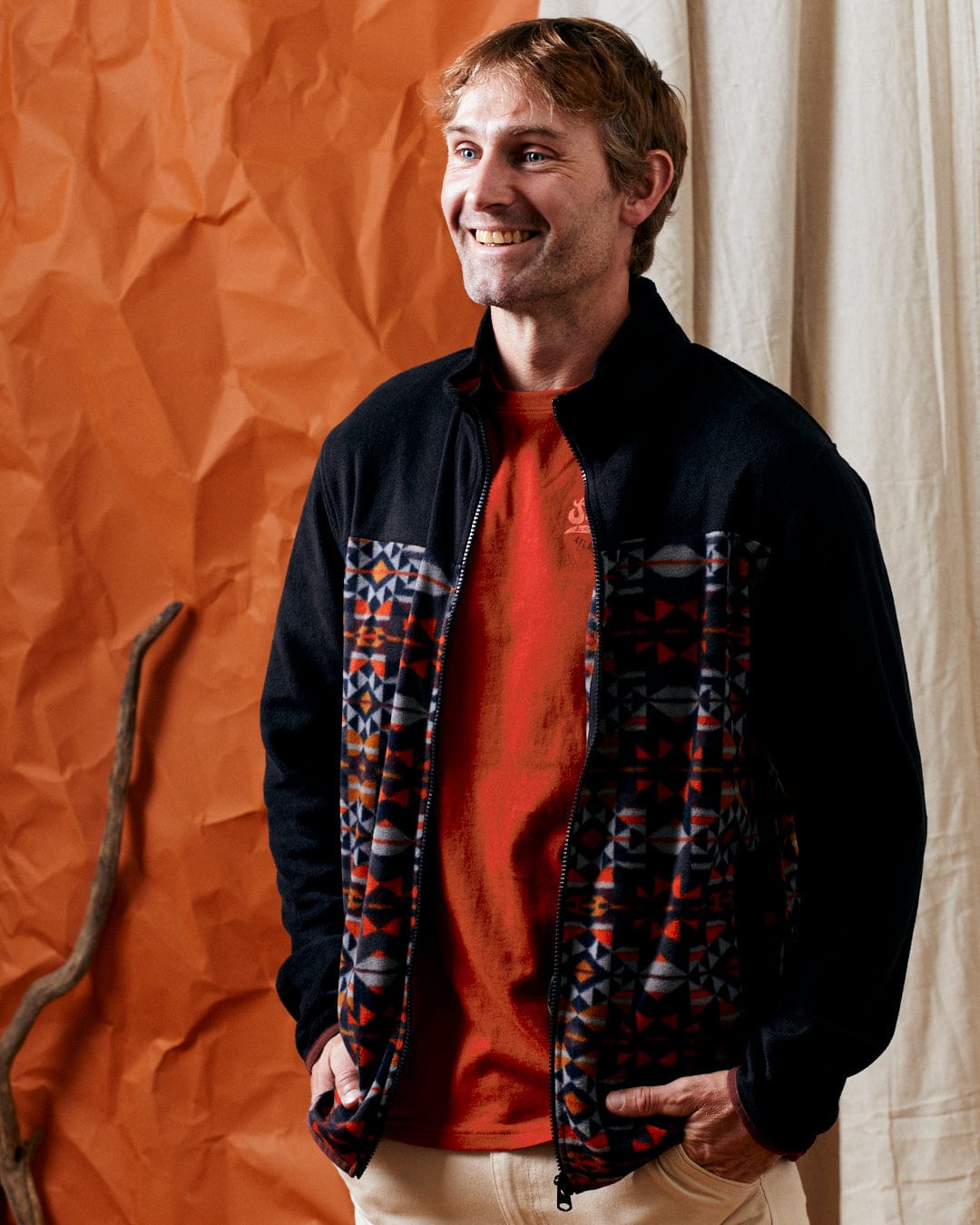 A man wearing the Theo Wilds Men's Recycled Microfleece jacket in black from Saltrock, over an orange shirt, stands smiling in front of an orange, crumpled backdrop with a curtain on one side and a stick leaning against the wall.