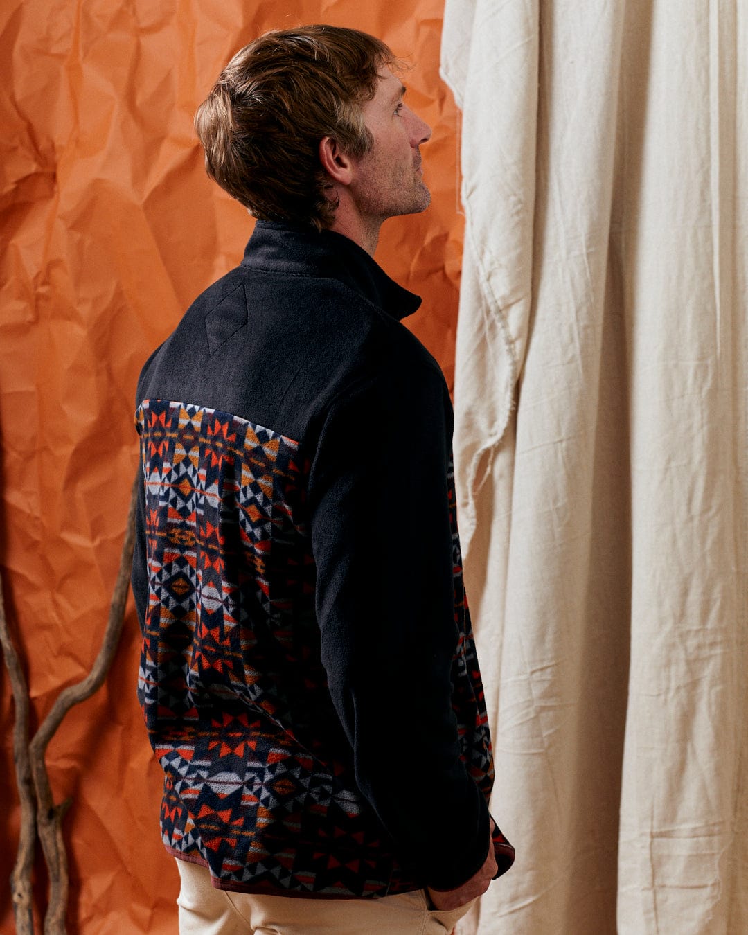 A person with short hair, wearing the Theo Wilds - Mens Recycled Microfleece in black by Saltrock, is looking up against a backdrop of crinkled orange paper and beige curtains.