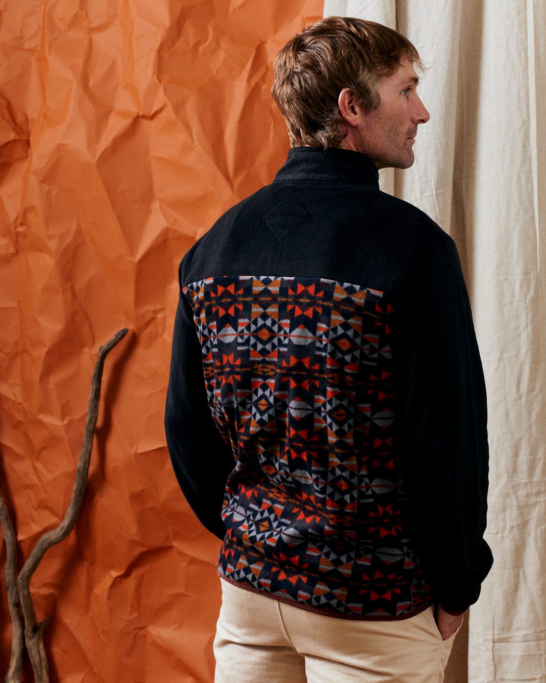 A person wearing the Saltrock Theo Wilds men's recycled microfleece jacket, which is black with a colorful geometric print on the back, stands sideways in front of a crumpled orange backdrop and a beige curtain, with a bare branch visible beside them.