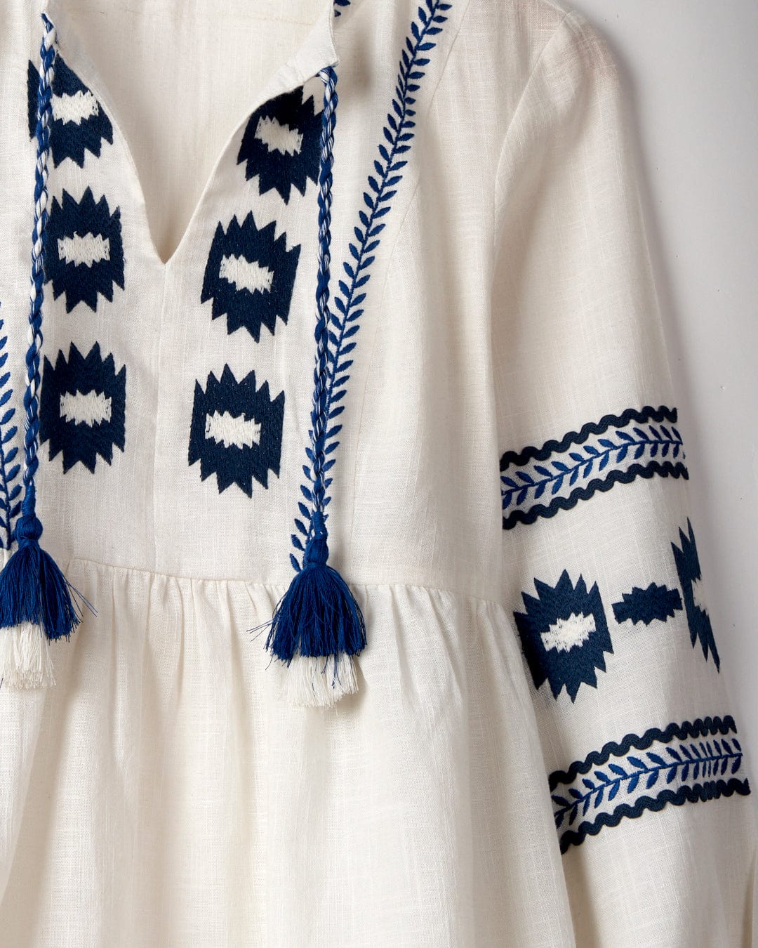 Close-up of the Tia - Women's Embroidered Blouse in white by Saltrock, made from 100% cotton with blue embroidered patterns. This smock-shaped garment features a v-neckline with tasseled cords and geometric designs on the chest and sleeves.