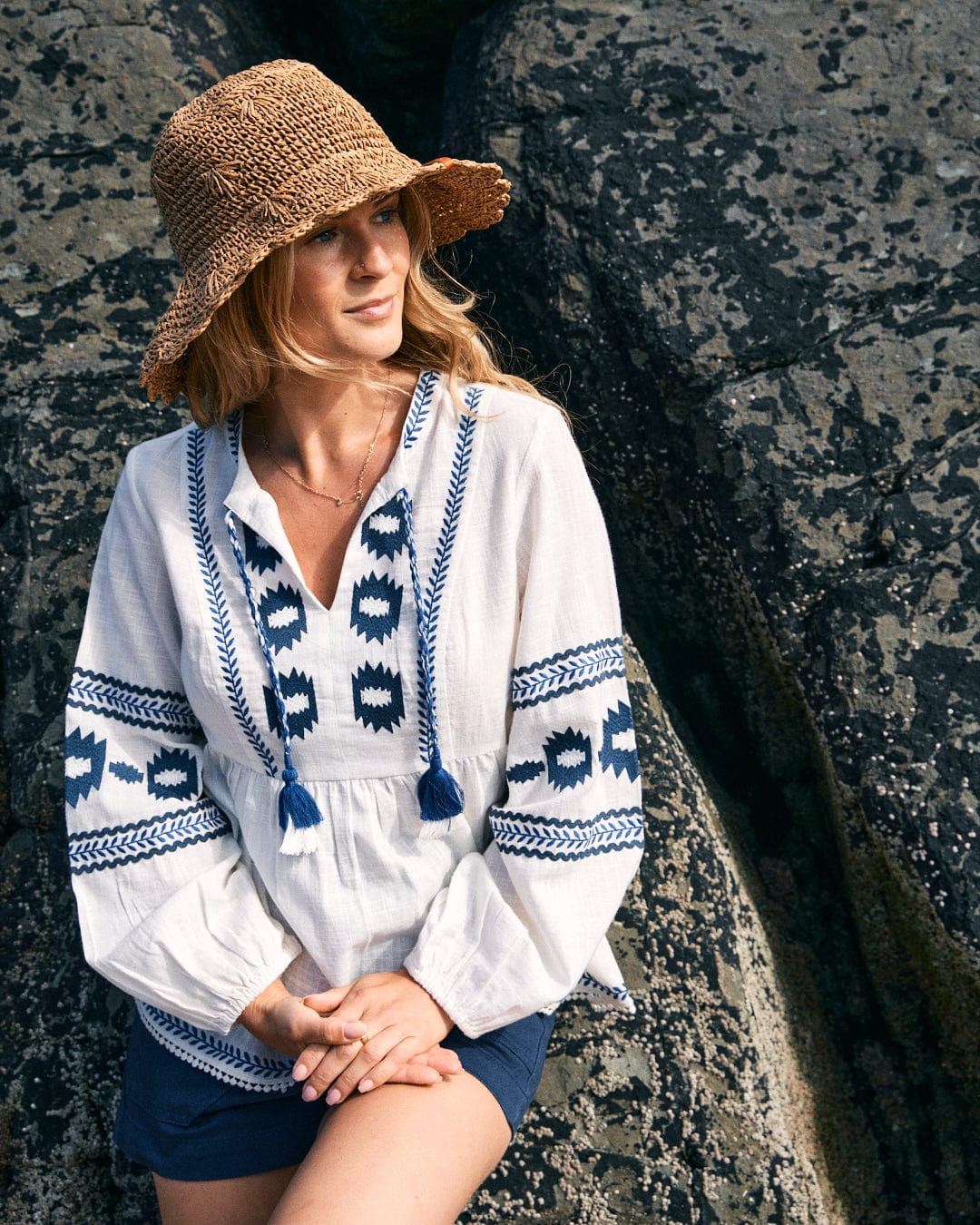 A woman wearing a straw hat, Saltrock's Tia - Womens Embroidered Blouse in white, and dark shorts sits against a rocky background. The blouse features intricate smock shape detailing and is made from 100% cotton.