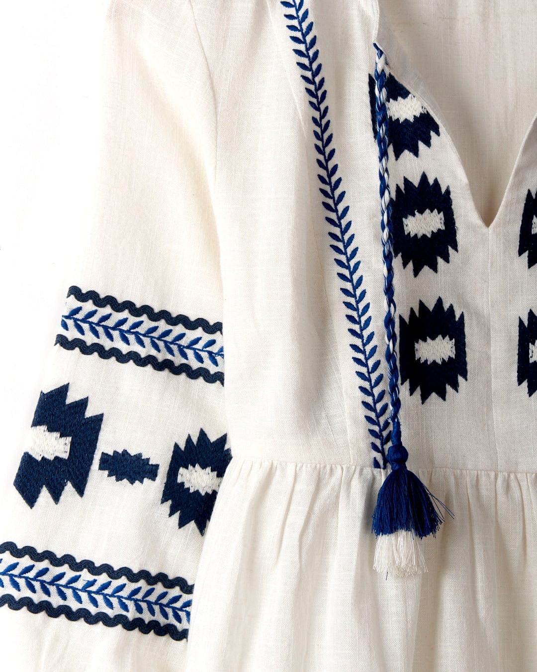 A close-up of the Tia - Womens Embroidered Blouse in white from Saltrock reveals intricate blue embroidered geometric patterns on the sleeves and chest, accented by braided tassel detailing. The smock shape lends a relaxed silhouette, while the 100% cotton fabric ensures ultimate comfort.