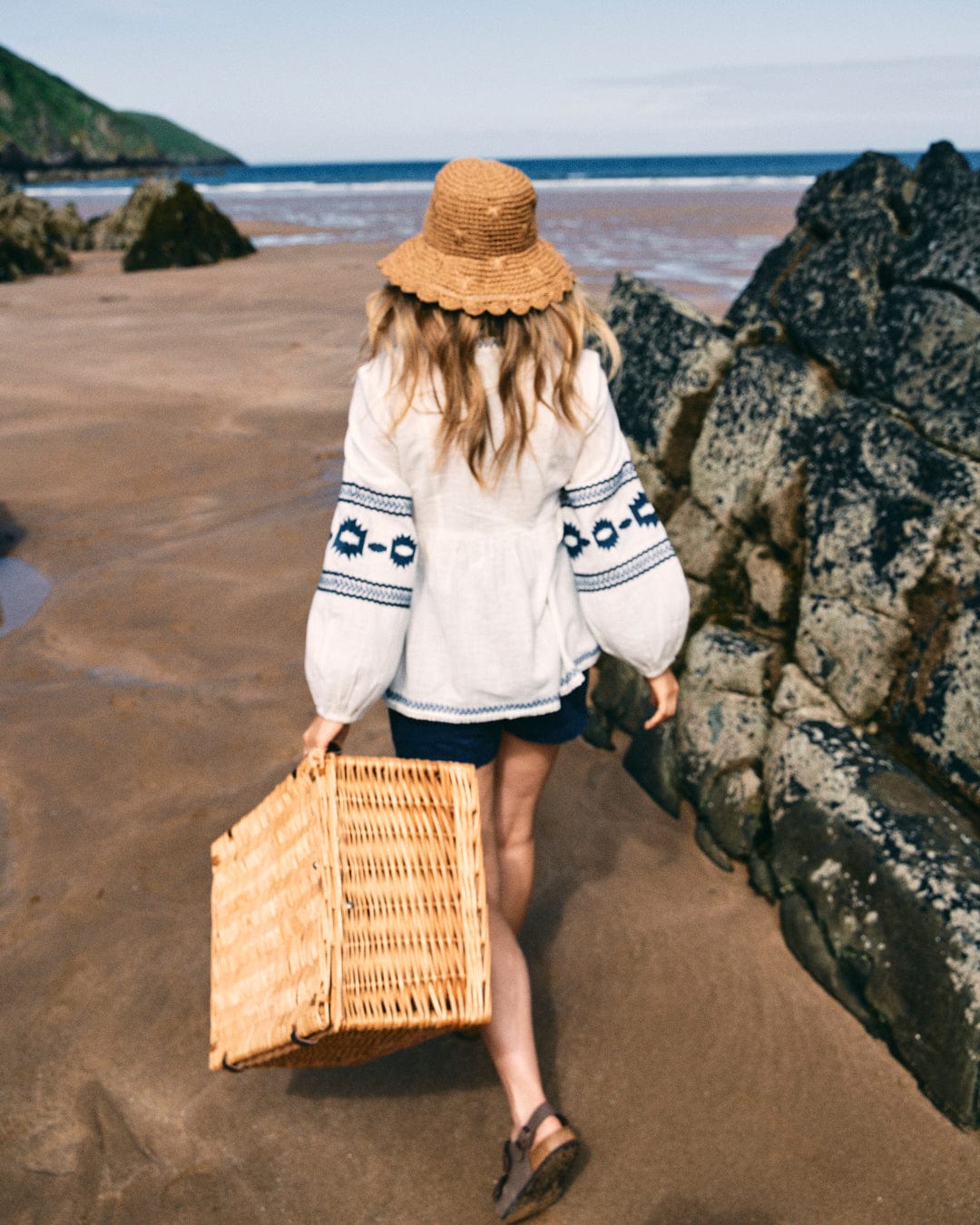 A woman wearing the Saltrock Tia Women's Embroidered Blouse in white, paired with a straw hat, walks along a sandy beach carrying a wicker basket. In the background, rocks and a lush green hill can be seen.