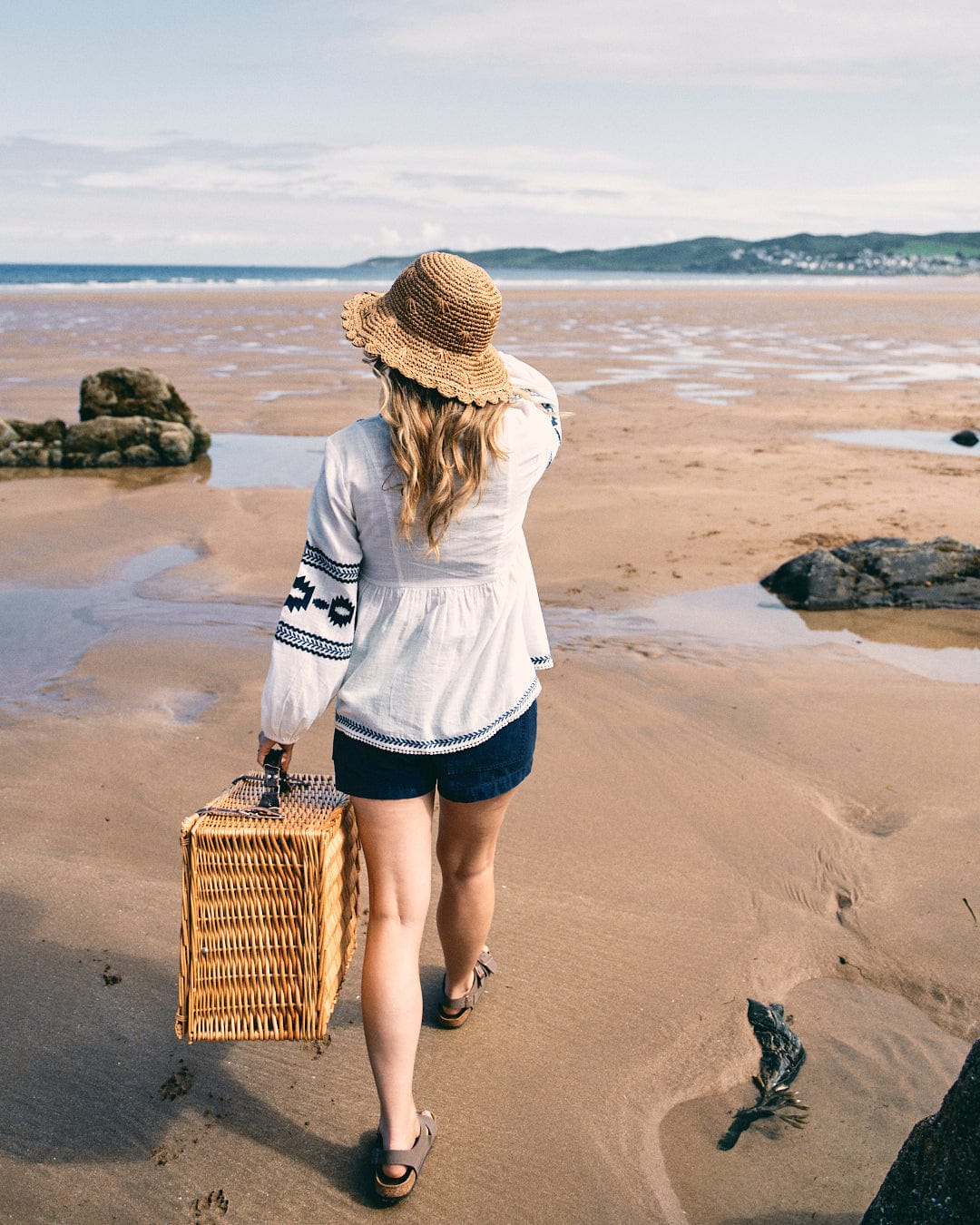 A woman in a Saltrock Tia blouse, featuring delicate embroidery on its white fabric, holds a wicker basket as she walks along a sandy beach toward the ocean.