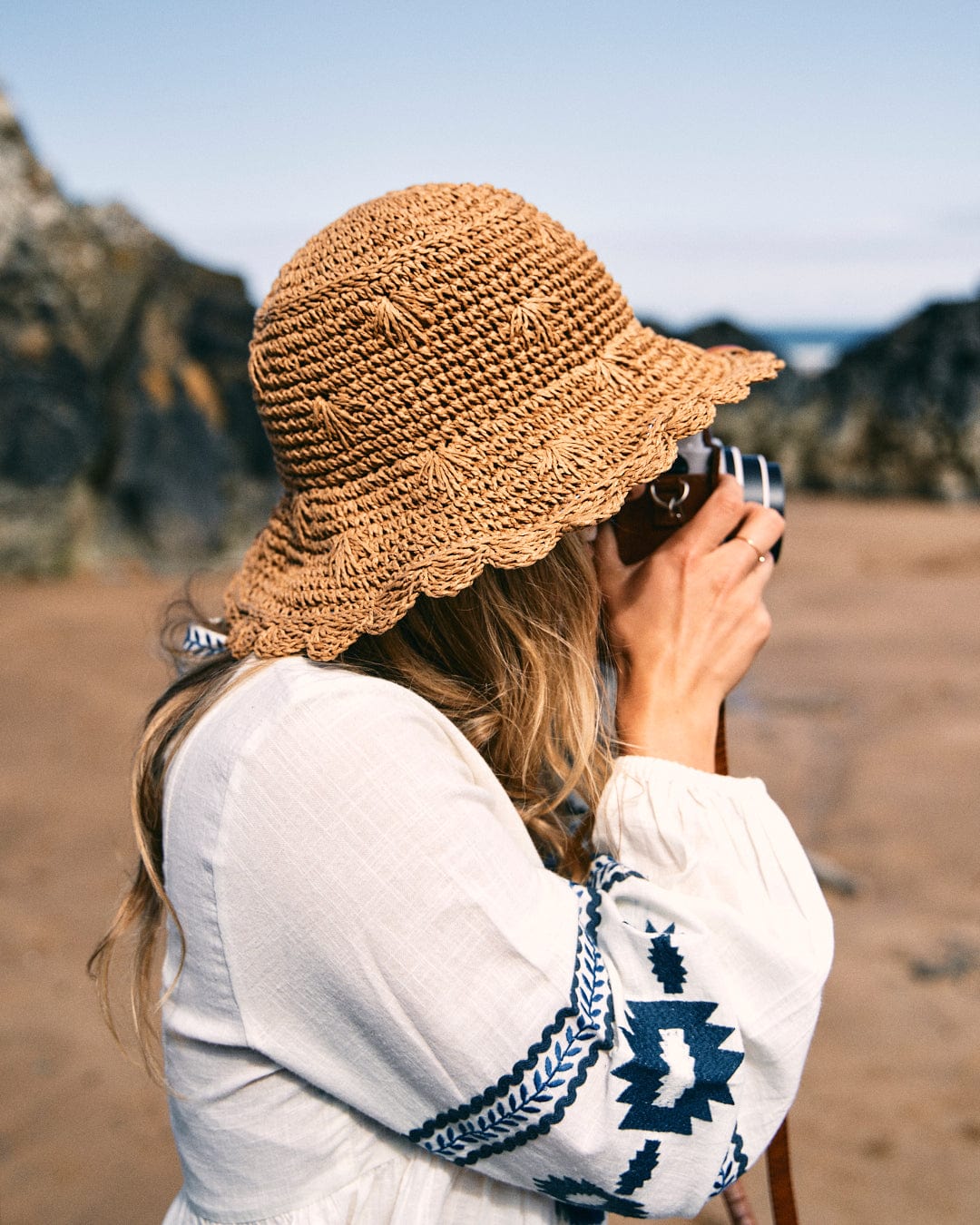 A person wearing a woven hat and the Saltrock Tia Women's Embroidered Blouse in white takes a photo with a camera on a rocky beach.