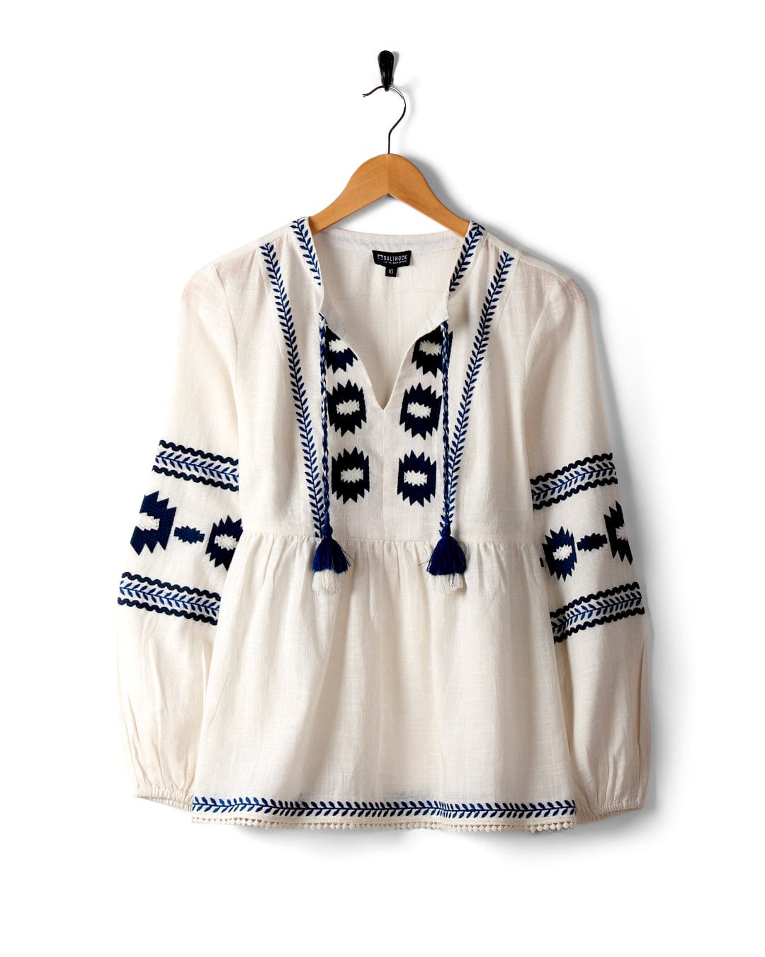 The Tia Women's Embroidered Blouse in white by Saltrock boasts blue geometric embroidery on the sleeves and front. Made from 100% cotton, it features a smock shape for optimal comfort and style. Displayed on a wooden hanger against a white background, this long-sleeve blouse is both stylish and versatile.