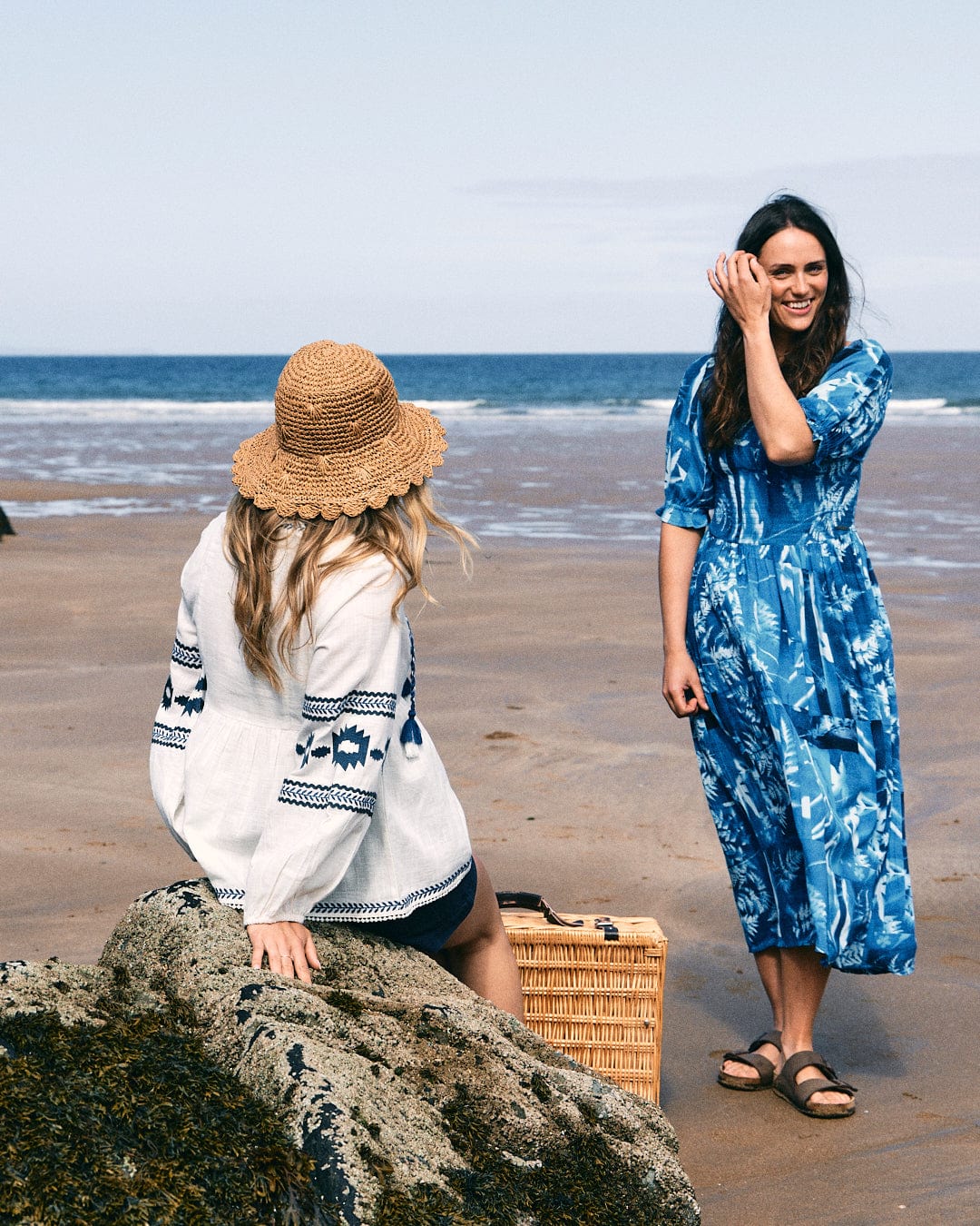 Two women are at the beach; one is sitting on a rock wearing a straw hat and the Saltrock Tia Women's Embroidered Blouse in white, made of 100% cotton, while the other stands nearby in a blue dress. A wicker basket is placed on the sand beside them.