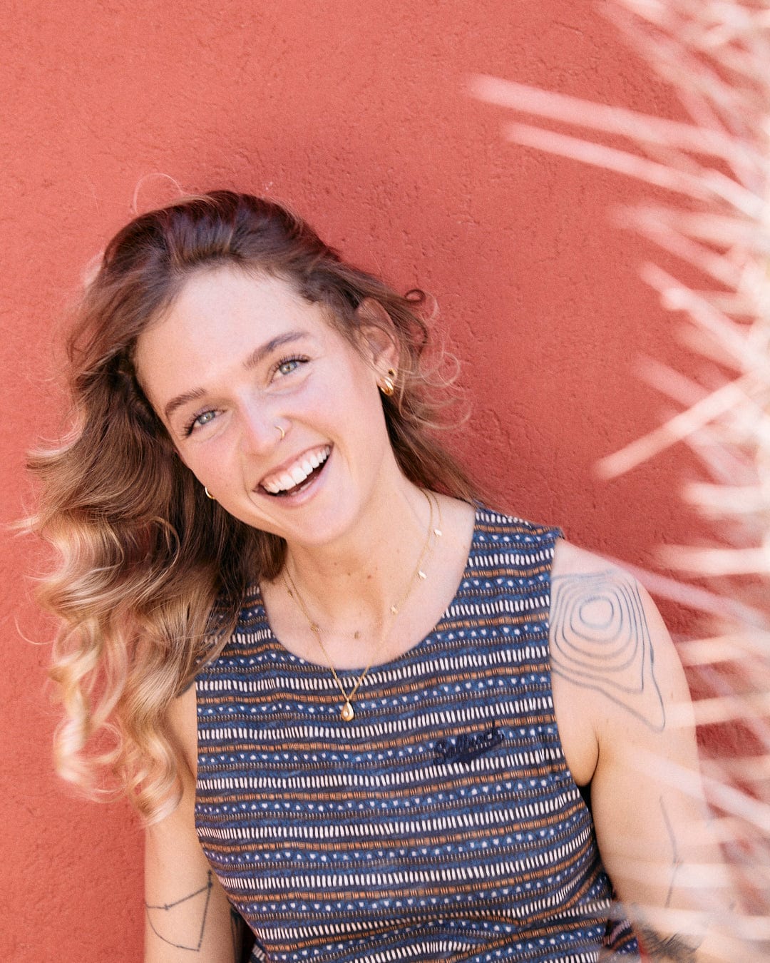 A woman with wavy hair smiles at the camera against a red wall background, wearing a Saltrock Tribal Stripe - Womens Midi Dress - Grey and displaying visible tattoos on her arm.