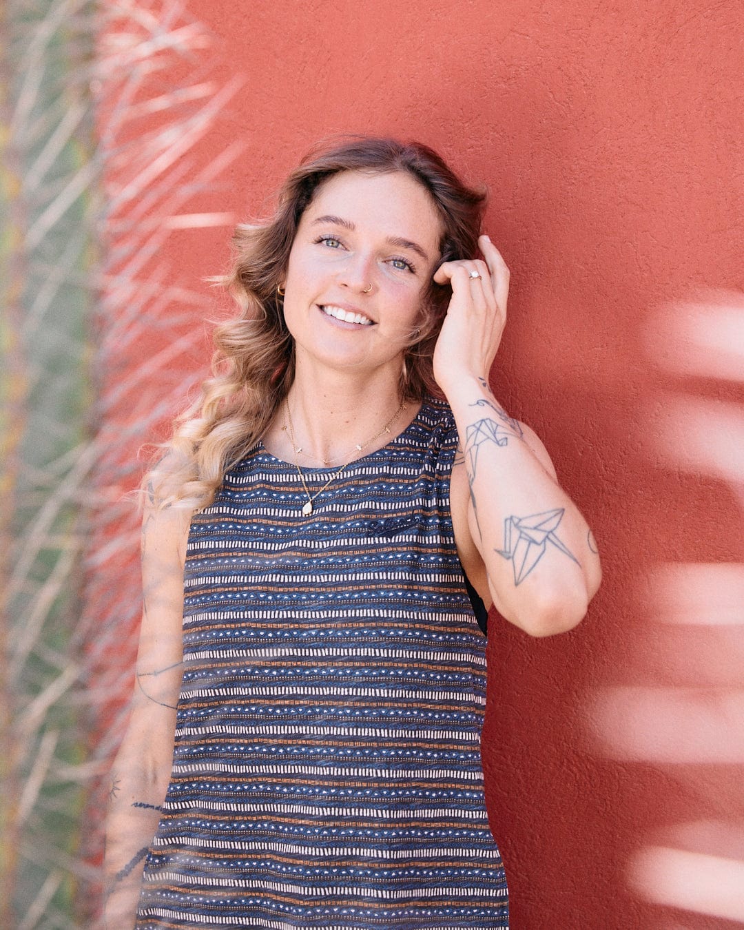 A woman with wavy hair and visible tattoos on her arm stands in front of a red wall, smiling and touching her hair. She is wearing the Saltrock Tribal Stripe - Womens Midi Dress - Grey featuring Saltrock branded embroidery.