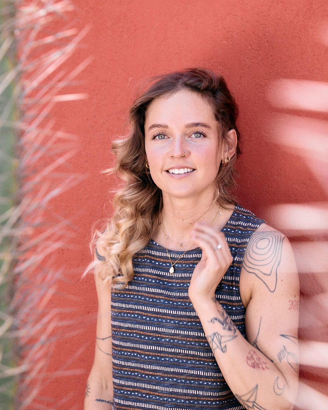 A person with wavy hair and tattoos on their arm, wearing a Saltrock Tribal Stripe - Womens Midi Dress - Grey, stands in front of a red wall with a neutral expression.