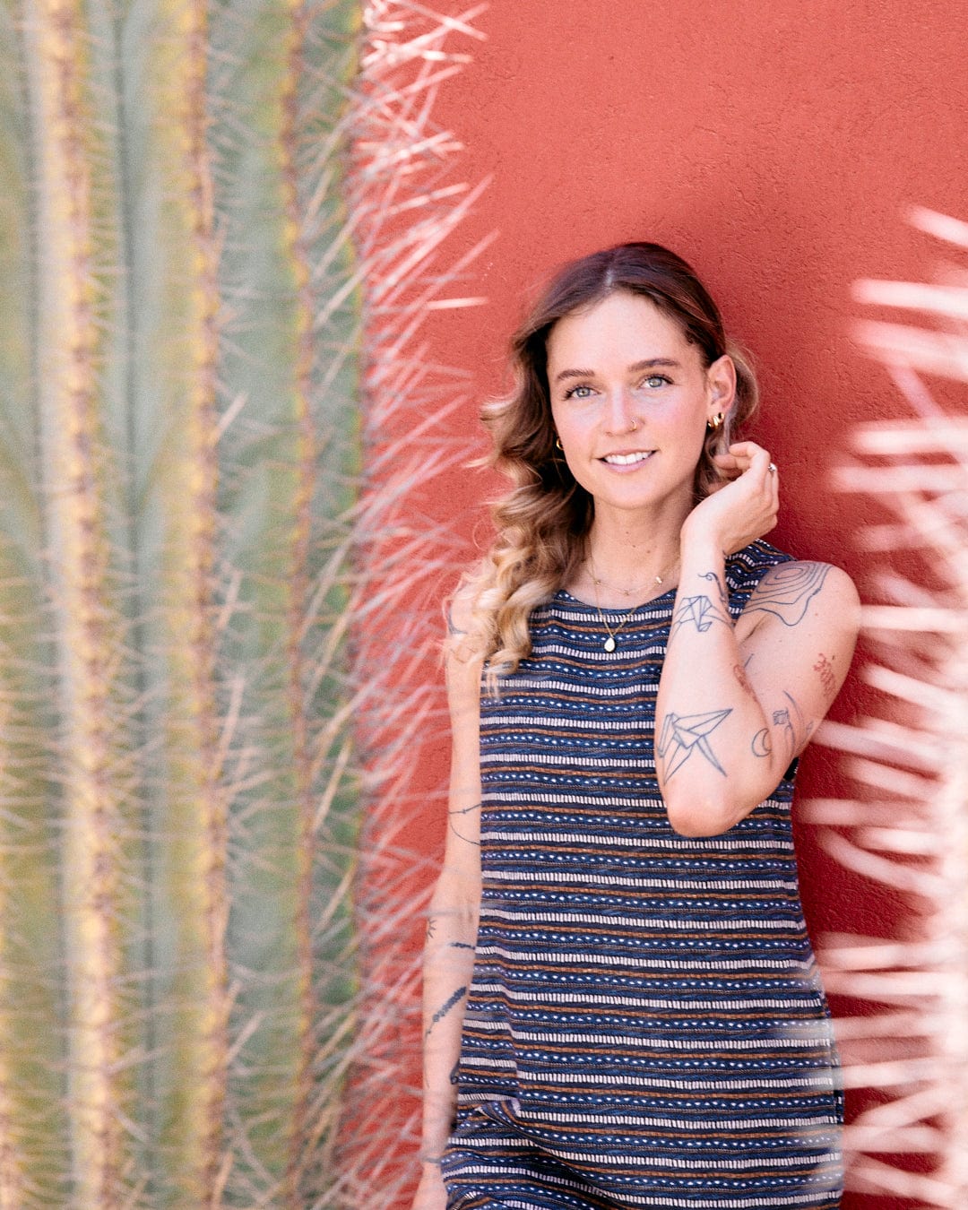 A woman with tattoos stands against a red wall beside a tall cactus. She is touching her hair and wearing a sleeveless Tribal Stripe - Womens Midi Dress - Grey with Saltrock branded embroidery.